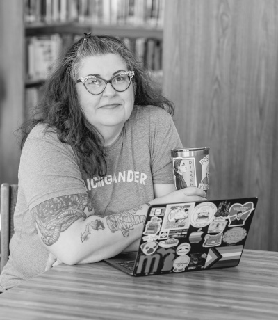 Black and white image of a woman sitting with her arms crossed. An open laptop is in front of her. She has long hair and is wearing glasses. Behind her is a bookshelf as she is in a library.